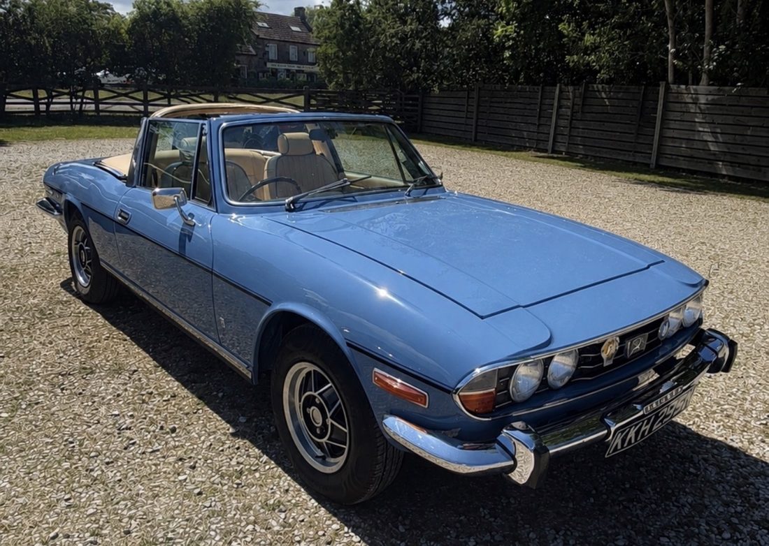 French Blue 1970s Triumph Stag convertible with tan interior, parked on gravel with the top down